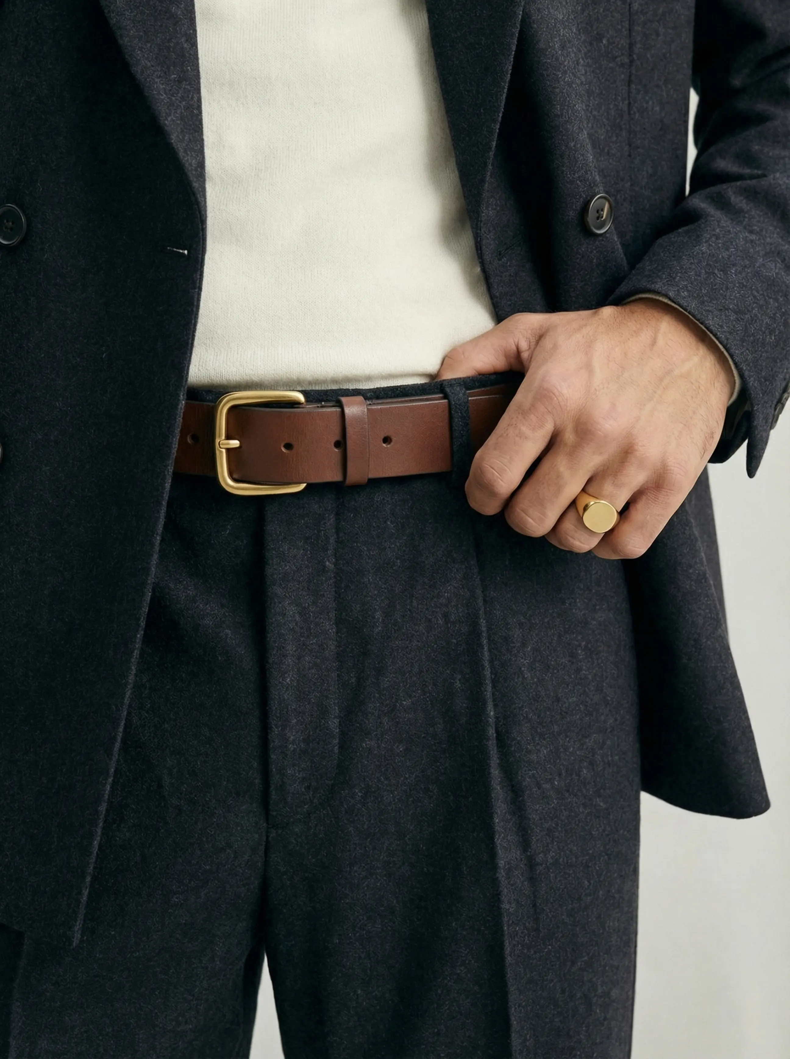Macro detail of belt buckle through trouser loops, textured wool lapel, and gold signet ring.