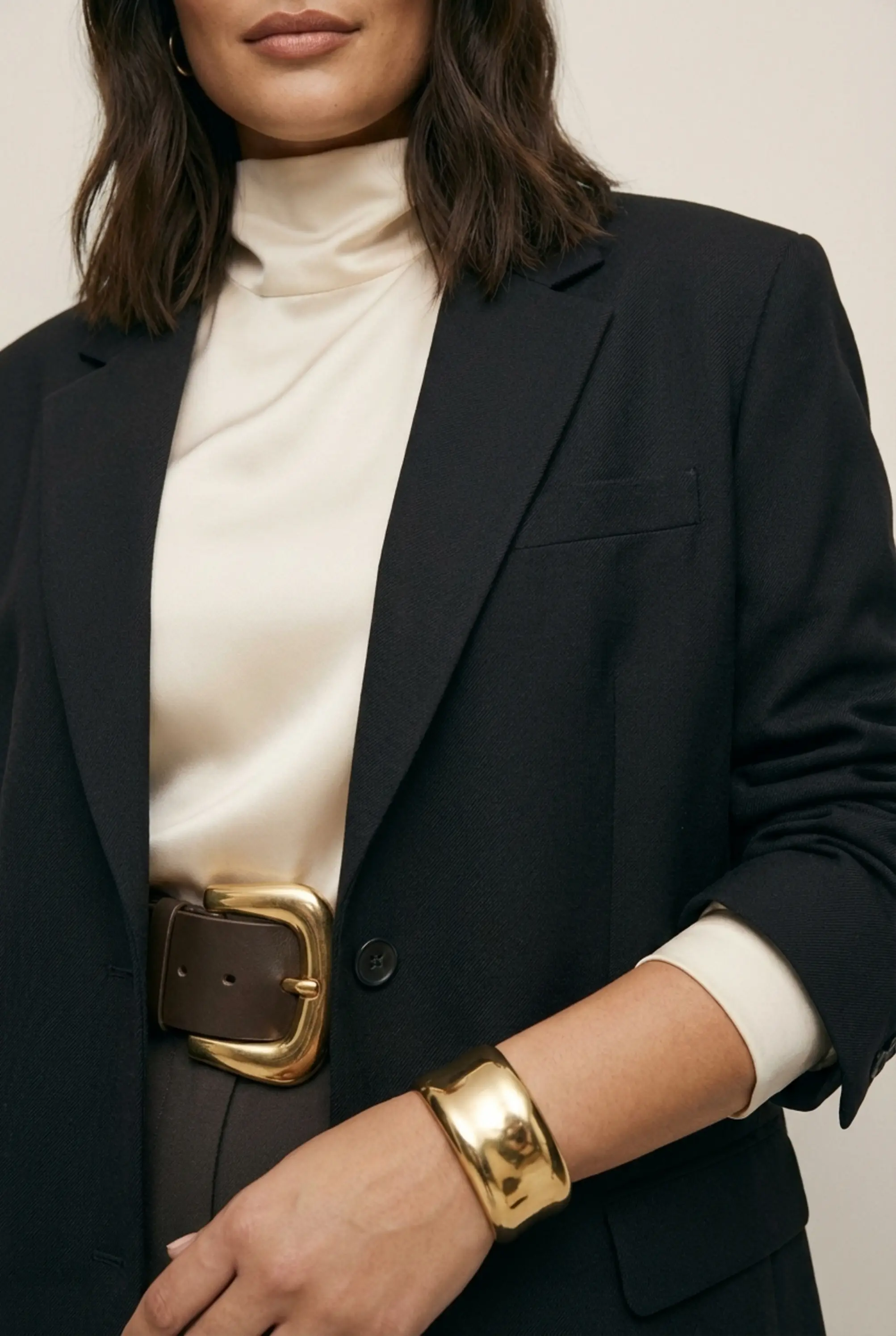 Macro detail of chunky belt buckle, textured wool blazer lapel and wide gold cuff with high-neck blouse.