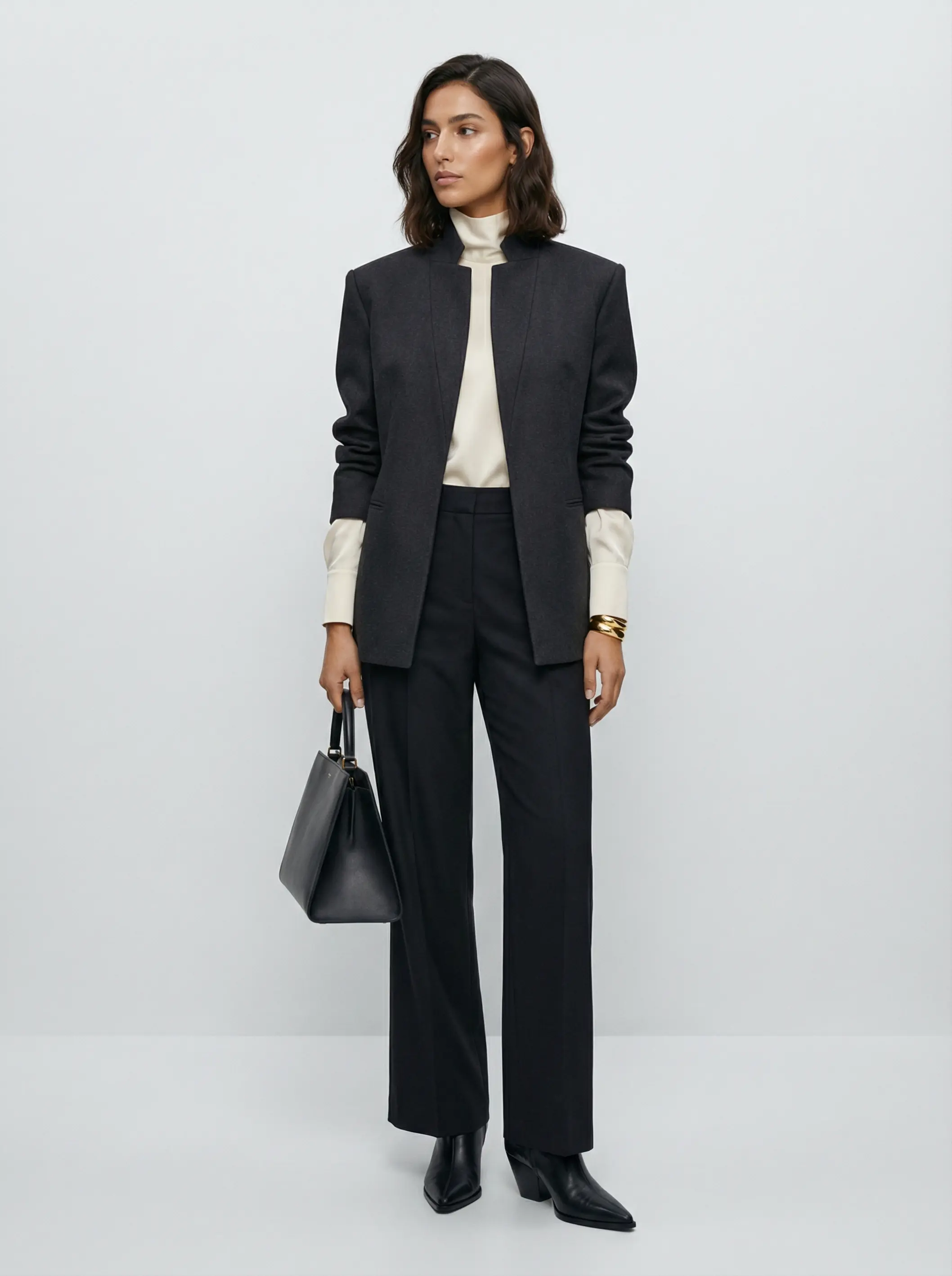 Full-body image of a woman in a high-collar sculpted jacket, wide-leg trousers and gold cuff on a neutral background.