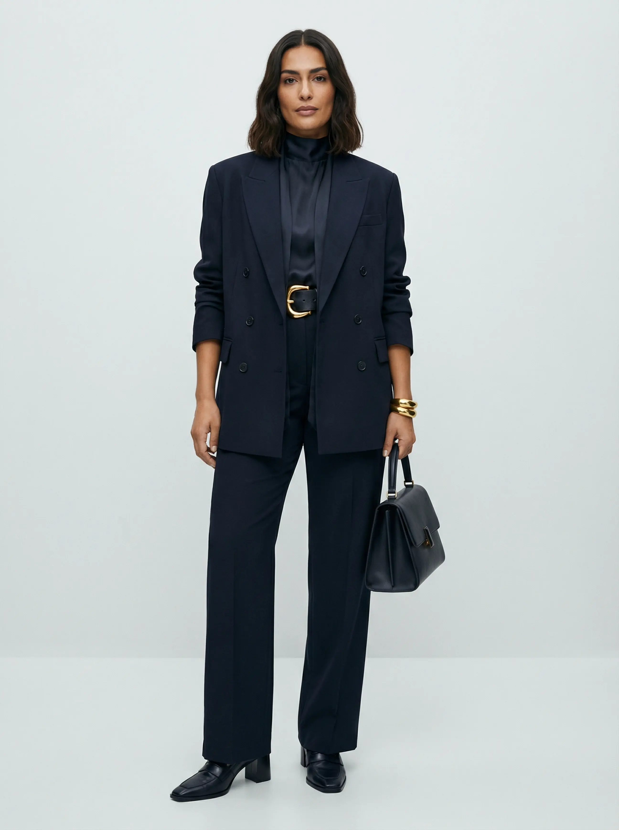 Full-body image of a woman in a baggy double-breasted blazer cinched with a chunky gold belt, wide-leg trousers, and gold cuff.