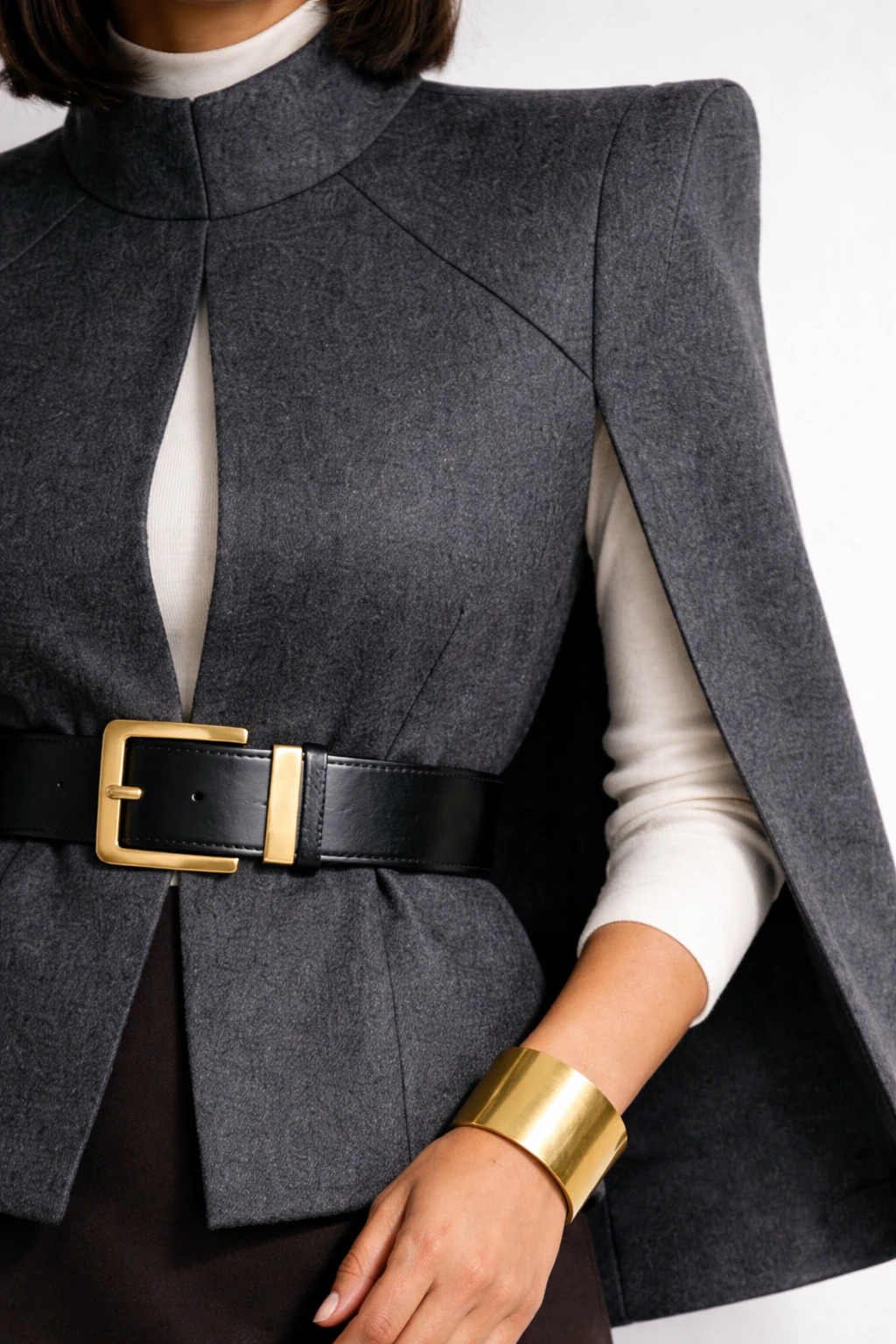 Close-up of brushed gold belt buckle cinching a charcoal cape blazer with an ivory mock-neck collar and a bold gold cuff bracelet.