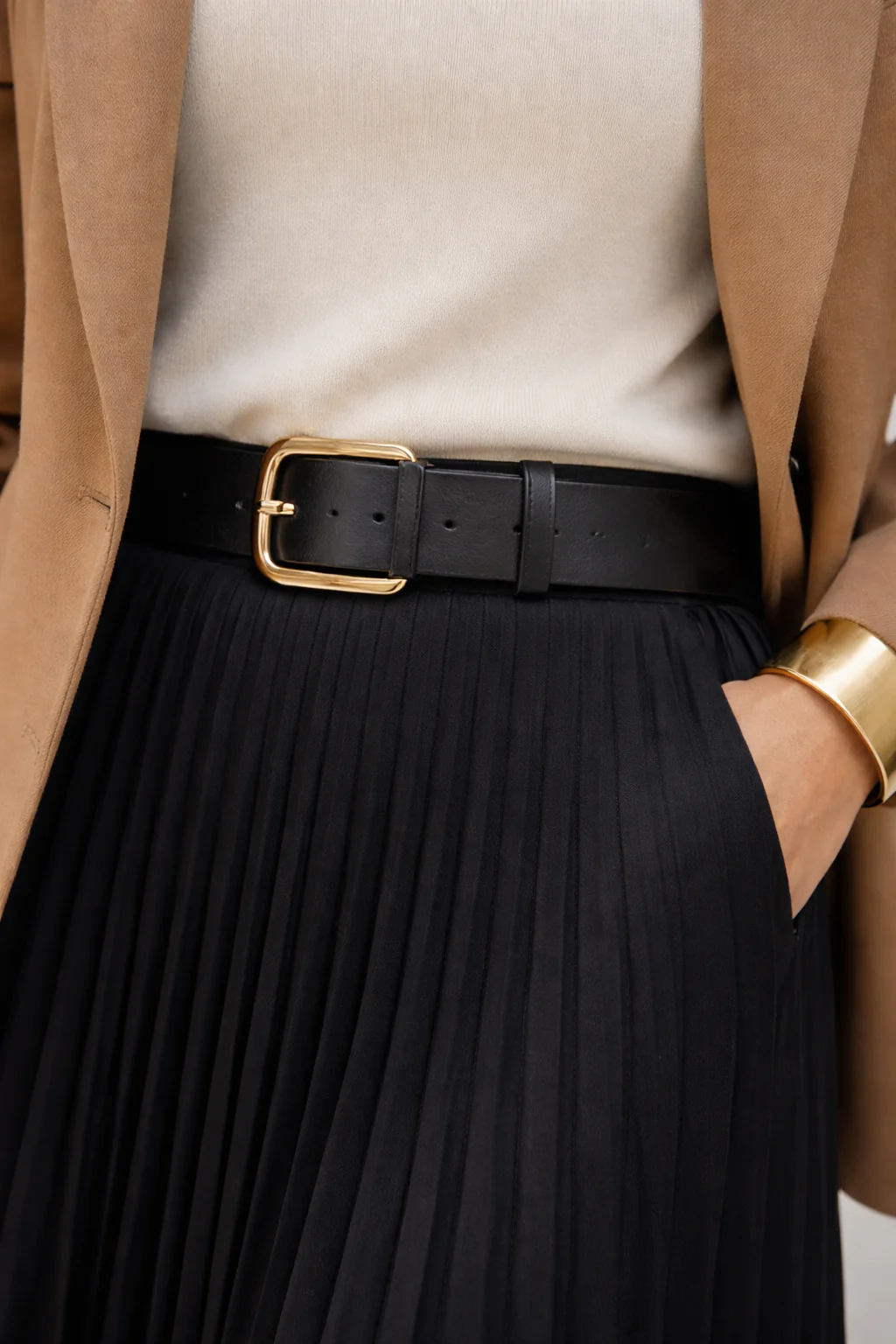 Macro close-up of a brushed-gold belt buckle over an ivory high-neck knit with black pleats and a bold gold cuff bracelet.